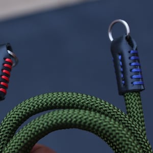 Puede incluir: Primer plano de una correa de cámara de cuerda verde con extremos de cuero negro. Los extremos de cuero están cosidos con hilo rojo y azul y sujetos a anillos metálicos. La correa está diseñada para una cámara.
