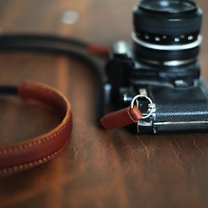 May include: A black camera with a brown leather strap. The camera has a silver ring around the lens.