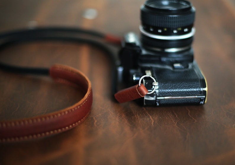 May include: A black camera with a brown leather strap. The camera has a silver ring around the lens.