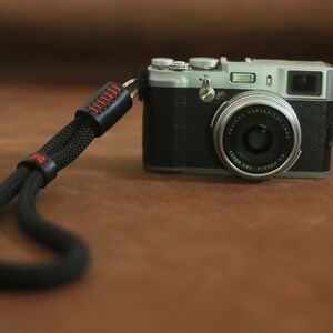 Può includere: Una fotocamera digitale argento e nera con un cinturino da polso in pelle nera. La fotocamera ha un obiettivo con il testo "FUJINON ASPHERICAL LENS SUPER EBC 14.2mm 1:2.8" stampato su di esso.