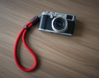 May include: A silver and black digital camera with a circular lens attached to a bright red rope wrist strap. The strap features black leather connectors with red stitching, and the camera body displays the Fujifilm brand name on the lens.
