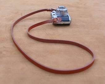 May include: A classic camera with a brown leather strap. The camera is silver and black, with a lens on top. The strap is thin and loops through a metal ring on the camera. The background is a light brown surface.