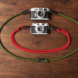 May include: Two vintage-style silver and black cameras, each attached to a rope camera strap. The top camera features a long olive green rope strap, while the bottom camera is connected to a shorter, vibrant red rope strap with black accents.
