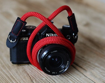 May include: A red camera strap made of braided rope with black leather ends wraps around a black camera with a silver lens. The lens has the text "Nikon" and "35mm 1:2.8 827155" printed on it.