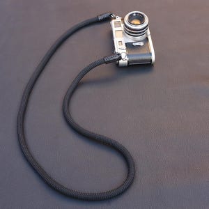 May include: A vintage silver camera with a black woven neck strap. The camera has a lens and a black leather grip. The strap is black and looped, resting on a dark surface.