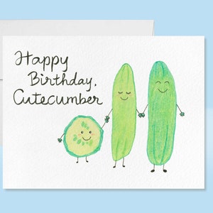 May include: A white card with the text "Happy Birthday, Cuecumber" written in black ink. Three cartoon cucumbers with faces are drawn on the card. The cucumbers are green and have black eyes and mouths. The smallest cucumber is a slice and the two larger cucumbers are holding hands.