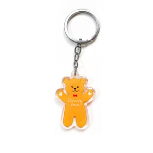 May include: A yellow teddy bear keychain with a red bow tie and the words "Bearing love" printed on its chest. The keychain is attached to a silver metal ring.