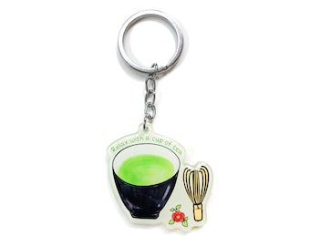 Könnte beinhalten: Ein weißer und grüner Schlüsselanhänger mit einer schwarzen Schale grünen Tees, einem Bambusbesen und einer roten Blume. Der Schlüsselanhänger trägt den Text "Relax with a cup of tea".