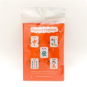 May include: A red card with the text "Mystery Keychain" and images of Mahjong tiles with cute faces. The text "Wishing you the best of luck!" and "sixtyeightcolors.com" are also on the card.