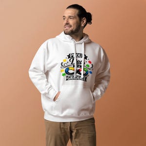 May include: A white hooded sweatshirt with a front pocket and a drawstring hood. The sweatshirt features a colorful graphic with the words "Proud Dad of a Graduate" and puzzle piece and infinity symbol designs.