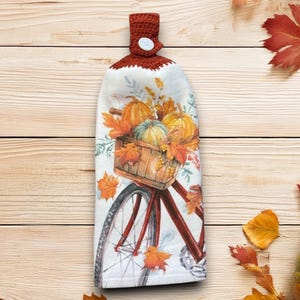 Puede incluir: Un paño de cocina colgante con temática otoñal. El paño presenta un diseño de acuarela de calabazas y hojas de otoño en una cesta sobre una bicicleta roja. La parte superior está tejida en ganchillo en color rojo oscuro con un botón blanco.