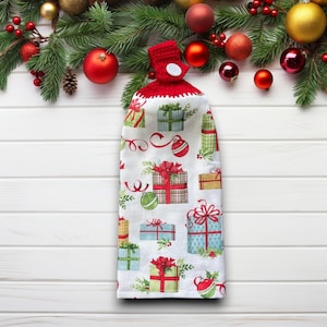 May include: A white kitchen towel with a red crocheted top and button closure. The towel is patterned with colorful gift boxes and Christmas ornaments. Festive holiday decor.