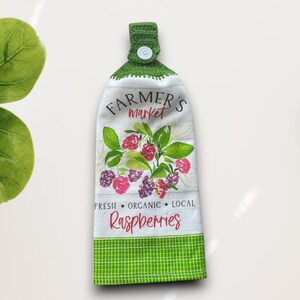 May include: White and green kitchen towel with a green crocheted loop and button. The towel features a watercolor design of raspberries and the text "Farmer's Market". The bottom border is green checkered.