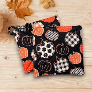 Puede incluir: Dos agarraderas negras con diseños de calabazas naranjas. Las calabazas tienen varios diseños, incluyendo lunares, rayas y cuadros. Cada agarradera tiene un lazo para colgar y un borde rojo.