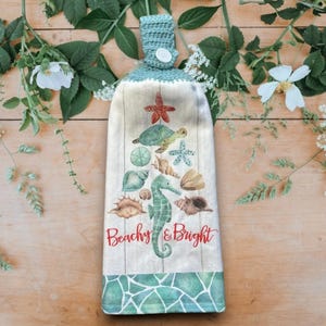 May include: A hanging kitchen towel with a teal crochet top and button. The towel features a beach-themed design with a seahorse, starfish, and seashells. The words "Beachy & Bright" are written in red script.