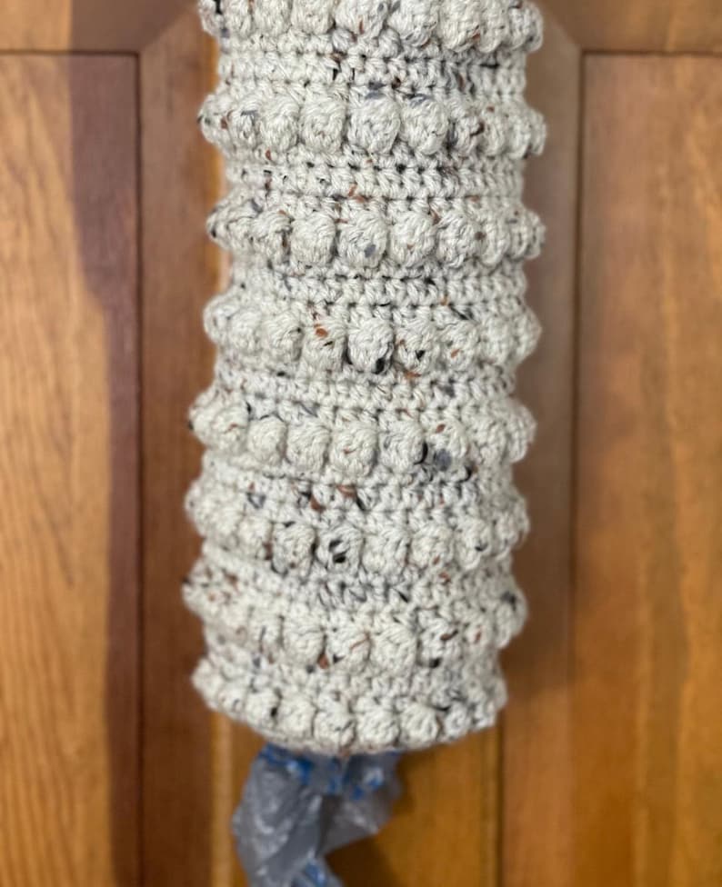 Modern boho style grocery bag holder. It measures 15 inches in length.  Features bobble crochet stitches in rows around it. Bag saver is hand crocheted in a beige fleck color. Large hook for hanging anywhere you like. Goes well with farmhouse style.
