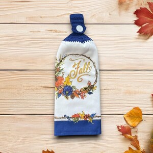 May include: Hanging kitchen towel in white and blue, with a fall theme. The towel has a wreath design with pumpkins, leaves, and the word "Fall" in gold script. The top is a dark blue crochet loop with a white button.