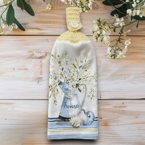 Pode incluir: Uma toalha de cozinha decorativa com um design em aquarela. A toalha apresenta um jarro azul cheio de flores brancas, a palavra "home" e um top de crochê amarelo com um botão. A parte inferior tem listras azuis e bege.