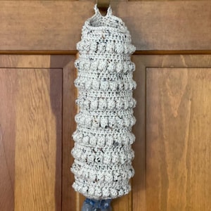 Modern boho style grocery bag holder. It measures 15" in length.  Features bobble crochet stitches in rows around it. Bag saver is hand crocheted in a beige fleck color. Large hook for hanging anywhere you like. Goes well with farmhouse style.