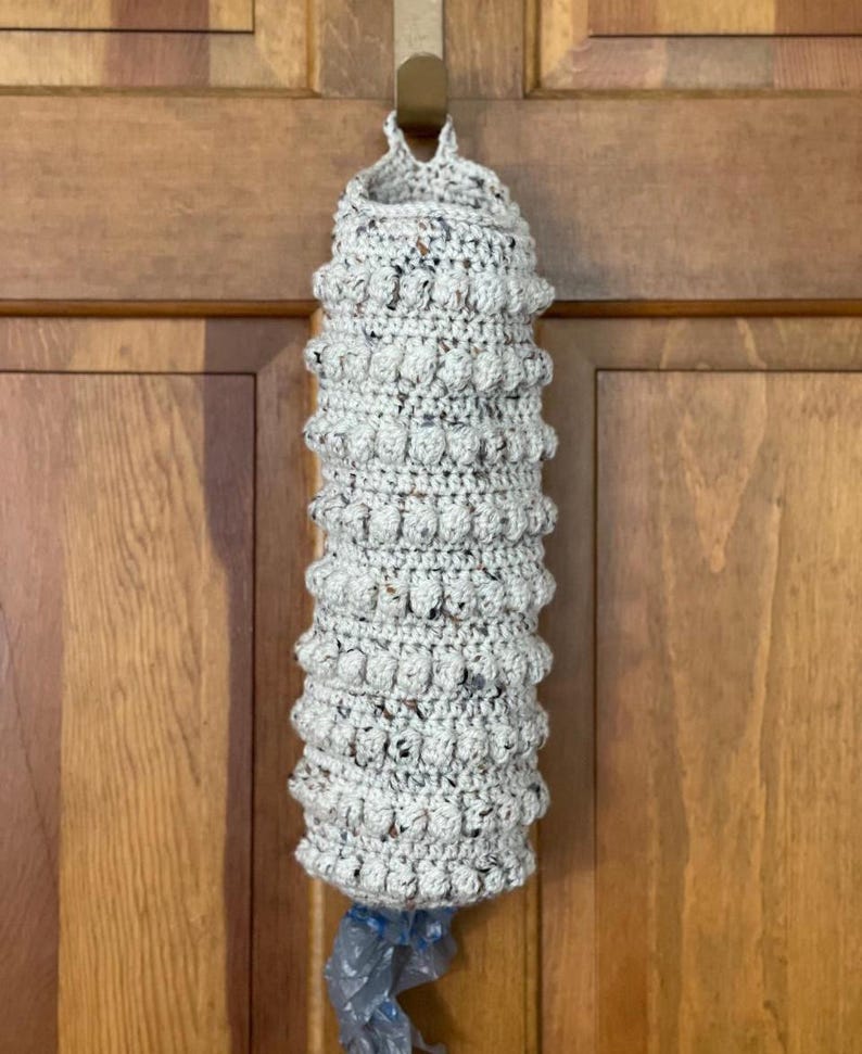 Modern boho style grocery bag holder. It measures 15" in length.  Features bobble crochet stitches in rows around it. Bag saver is hand crocheted in a beige fleck color. Large hook for hanging anywhere you like. Goes well with farmhouse style.