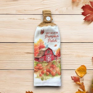 May include: A white kitchen towel with a brown crocheted top and button closure. The towel features a watercolor illustration of a red barn, pumpkins, and fall foliage, with the text "Meet Me At The Pumpkin Patch".