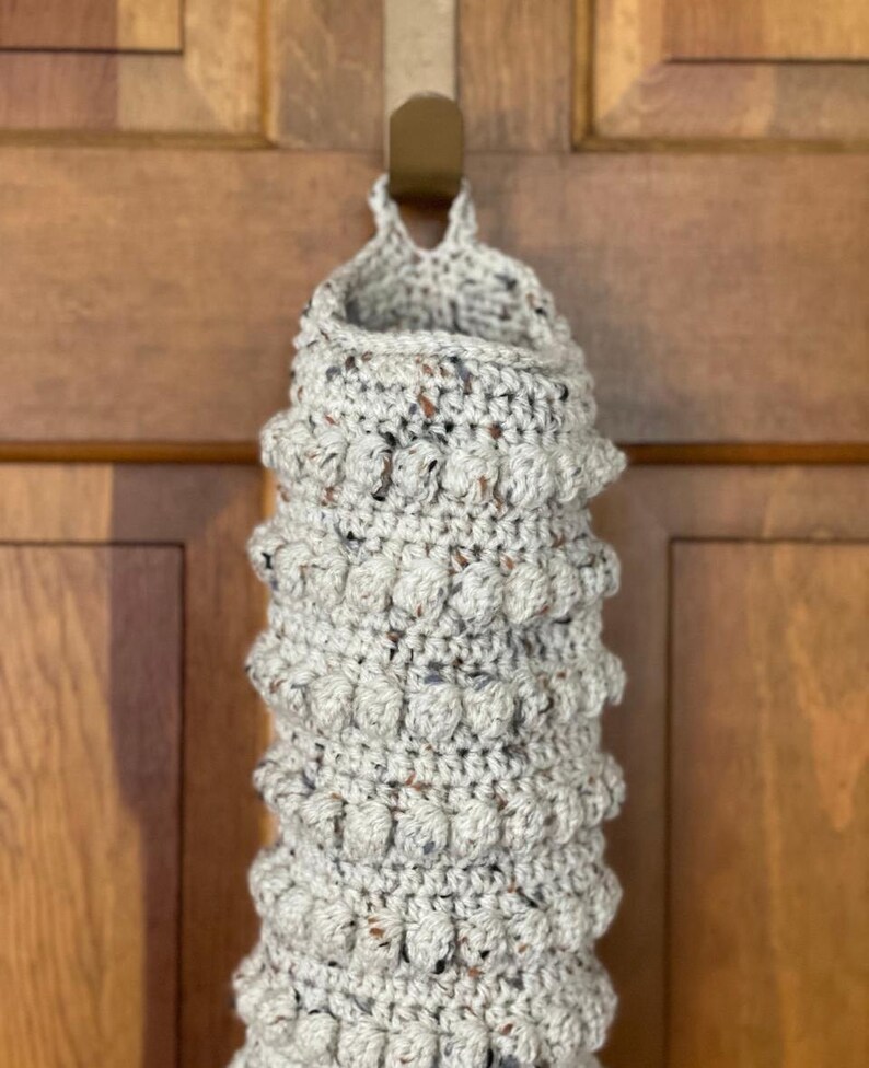 Modern boho style grocery bag holder. It measures 15 inches in length.  Features bobble crochet stitches in rows around it. Bag saver is hand crocheted in a beige fleck color. Large hook for hanging anywhere you like. Goes well with farmhouse style.