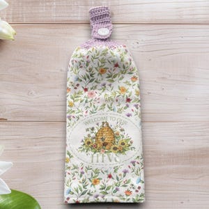 Puede incluir: Un paño de cocina colgante con un estampado floral y una parte superior de ganchillo. La toalla presenta un diseño ovalado central con una ilustración de colmena y el texto "WELCOME TO OUR HIVE". El estampado floral incluye varios colores.