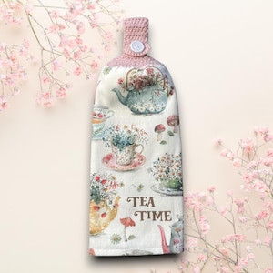May include: A pink and white tea towel with a floral pattern and teacups. The towel has a pink crocheted loop and a button. The text "TEA TIME" is printed on the towel.