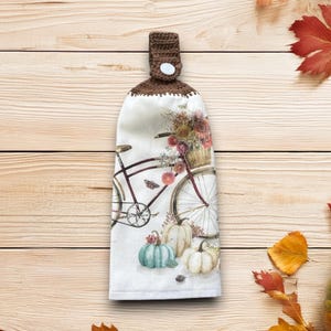 May include: A hanging kitchen towel with a brown crocheted top and a button closure. The towel features a watercolor design of a bicycle with a basket of flowers, pumpkins, and fall leaves.