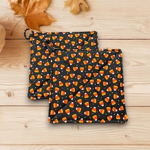 May include: Two black square pot holders with a candy corn pattern. The candy corn is orange, yellow, and white. The pot holders are on a wooden surface with autumn leaves and a white pumpkin.