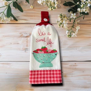 May include: A white hanging kitchen towel with a red crocheted top featuring a white button. The towel displays a teal colander filled with red strawberries and the red text It's a sweet life. The bottom edge has a red and white checkered pattern.