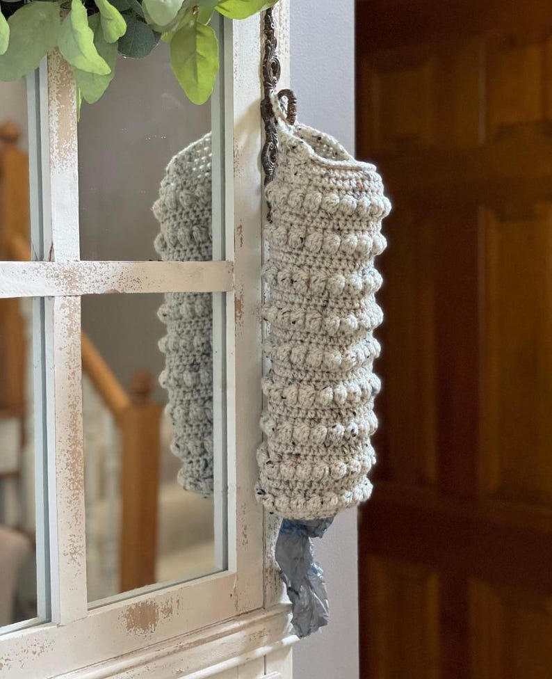 Modern boho style grocery bag holder. It measures 15 inches in length.  Features bobble crochet stitches in rows around it. Bag saver is hand crocheted in a beige fleck color. Large hook for hanging anywhere you like. Goes well with farmhouse style.