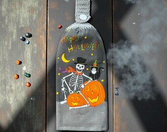 Peut inclure: Essuie-mains de cuisine gris pour Halloween avec un dessus en crochet. Le torchon présente un squelette, un chat noir et des citrouilles avec l'inscription "Happy Halloween". Le torchon est suspendu sur une surface en bois.