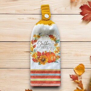 Può includere: Un canovaccio da cucina appeso con una parte superiore all'uncinetto gialla e una chiusura con bottone. L'asciugamano bianco presenta un design a tema autunnale con una zucca, girasoli e le parole "Gather Here". Il fondo ha strisce arancioni e verde acqua.