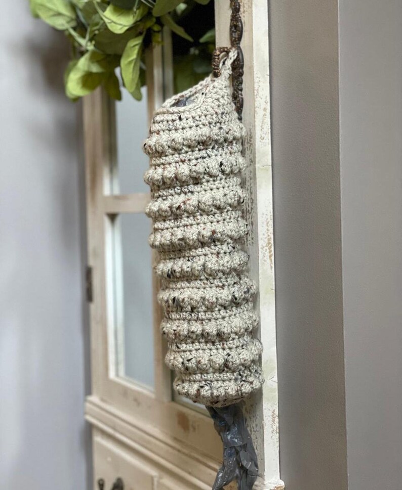 Modern boho style grocery bag holder. It measures 15 inches in length.  Features bobble crochet stitches in rows around it. Bag saver is hand crocheted in a beige fleck color. Large hook for hanging anywhere you like. Goes well with farmhouse style.