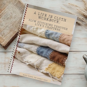 Op de afbeelding: Een spiraalgebonden werkboek getiteld A LIFE IN CLOTH WORKBOOK: SLOW STITCHING THROUGH LIFE'S CHAPTERS door ALICE DOWN THE RABBIT HOLE. De omslag toont een foto van gelaagde, met de hand gestikte stoffen lappen in crème, blauw en bruin.