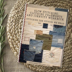 Op de afbeelding: Een spiraalgebonden werkboek getiteld SLOW STITCHING: CLOTHING FRAGMENTS AND IDENTITY WORKBOOK STITCHING THE STORIES WE WEAR door Alice Down The Rabbit Hole. De kaft toont een collage van stoffen lapjes, inclusief spijkerstof zakken en borduurwerk.