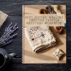 Könnte beinhalten: Ein Arbeitsbuch mit dem Titel "Soul Cloths: Healing Through Textile Gestures Workbook" mit Spiralbindung. Das Cover zeigt ein genähtes Tuch und Stickgarn. Eine schwarze Teekanne, Lavendel und Jute sind ebenfalls vorhanden.