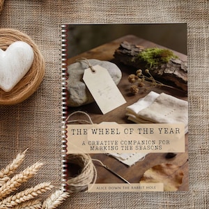Könnte beinhalten: Ein Spiralbuch mit dem Titel "THE WHEEL OF THE YEAR" und dem Untertitel "A CREATIVE COMPANION FOR MARKING THE SEASONS". Das Bild zeigt ein weißes Herz, Bindfaden, Weizenähren und ein Etikett. Das Buch liegt auf einer rustikalen, strukturierten Oberfläche.
