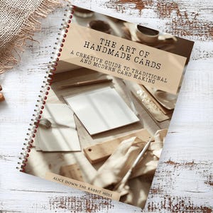 May include: A spiral-bound book titled "The Art of Handmade Cards" with the subtitle "A Creative Guide to Traditional and Modern Card Making." The cover features a sepia-toned image of card-making supplies. Cinnamon sticks, sprigs of greenery, and burlap are also visible.