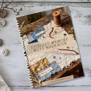 Könnte beinhalten: Ein spiralgebundenes Arbeitsbuch mit dem Titel "Stitched Memory Maps Workbook" mit rustikalem Design. Das Cover zeigt bestickte Stoffflicken, eine Holzspule mit Faden und den Text "Alice Down the Rabbit Hole". Dekorative Knöpfe und Spitze sind um das Buch angeordnet.