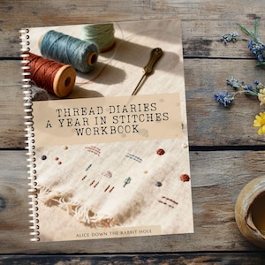 May include: A workbook titled "Thread Diaries: A Year in Stitches" with spools of thread, a needle, and a cup of tea. The book is open, showing embroidery. A bouquet of wildflowers sits nearby on a wooden surface.