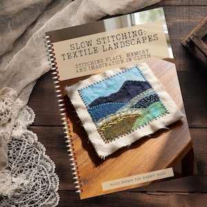 Könnte beinhalten: Ein Buch mit dem Titel "SLOW STITCHING: TEXTILE LANDSCAPES" mit einem gestickten Landschaftsbild auf dem Einband. Das Buch ist geöffnet und zeigt den Titel und den Untertitel. Der Einband zeigt eine gestickte Landschaft mit blauen, grünen und braunen Farbtönen. Baumwollkugeln und ein Holztablett sind im Hintergrund.