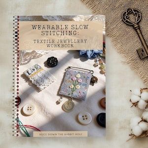 May include: A spiral-bound workbook titled "Wearable Slow Stitching: Textile Jewellery Workbook" with various craft supplies. The image includes a vintage key, cotton sprigs, buttons, and embroidered textile jewelry.