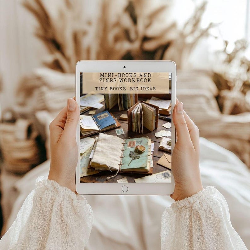May include: A tablet displaying a digital workbook titled MINI-BOOKS AND ZINES WORKBOOK, TINY BOOKS, BIG IDEAS. The screen shows an arrangement of small handmade books, loose papers, and a dried flower on a dark wooden surface.