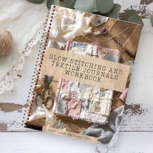 May include: A spiral-bound workbook titled "SLOW STITCHING AND TEXTILE JOURNALS WORKBOOK" with a collage cover. The cover features fabric scraps, scissors, and a ball of twine. The book is on a distressed white wooden surface.
