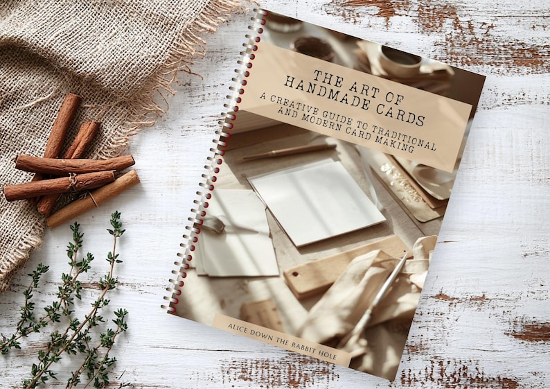 Pu&ograve; includere: Un libro con rilegatura a spirale intitolato The Art of Handmade Cards: A Creative Guide to Traditional and Modern Card Making di Alice Down the Rabbit Hole. La copertina presenta una fotografia di cancelleria, una penna e un blocco di legno.