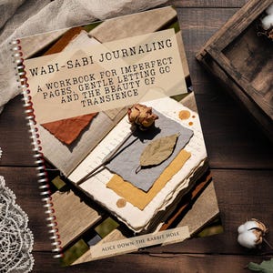 Könnte beinhalten: Ein Spiralbuch mit dem Titel "Wabi-Sabi Journaling" mit dem Text "A Workbook for Imperfect Pages, Gentle Letting Go and the Beauty of Transience." Das Journal zeigt eine Collage aus strukturierten Papieren und eine getrocknete Rose. Eine Holzkiste mit Baumwollknospen befindet sich im Hintergrund.