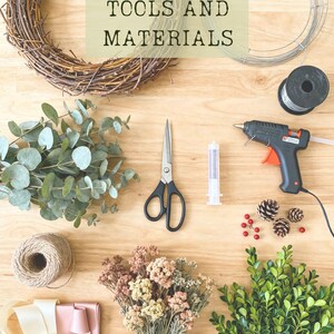 K&ouml;nnte beinhalten: Eine Sammlung von Bastelmaterialien, darunter ein brauner Zweigkranz, ein silberner Drahtrahmen, eine Drahtspule, eine Hei&szlig;klebepistole, eine Schere, eine Spritze, Bindfaden, cremefarbene und rosa B&auml;nder, Eukalyptus, Trockenblumen, Tannenzapfen und Buchsbaum.