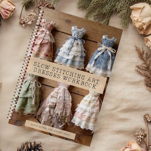 Può includere: Un quaderno a spirale intitolato "Slow Stitching Art Dresses Workbook" con abiti in miniatura di tessuto di vari colori. La copertina è su una superficie di legno, circondata da fiori secchi e pigne.